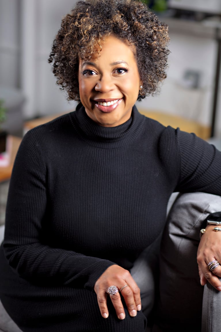 Portrait of a smiling woman with curly brown hair wearing a black turtleneck sweater, seated indoors